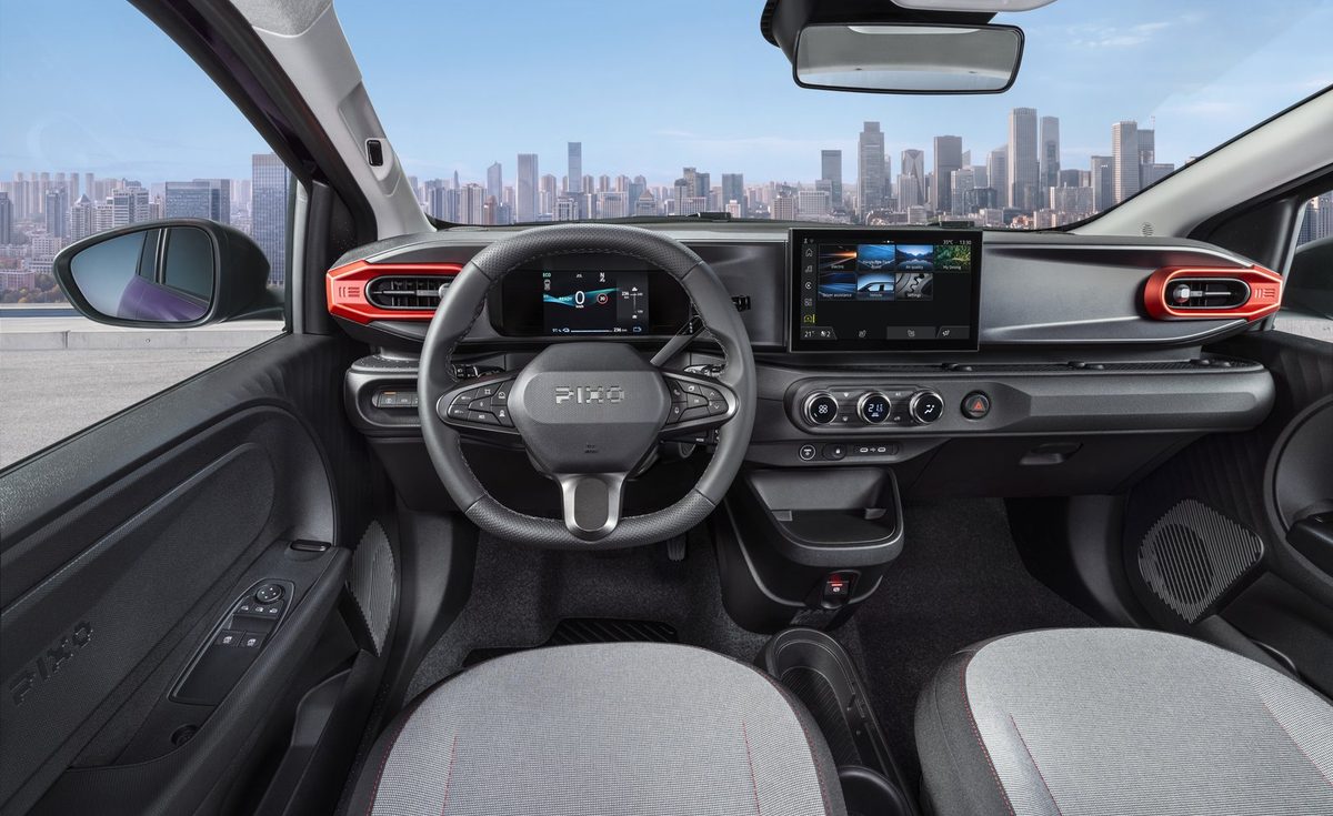 Photograph showcasing the all-new PIXO interior with steering wheel, digital instrument cluster, central touchscreen, climate controls, and textured seats. Red-accented air vents, black dashboard trim, compact center console, and city skyline visible through windshield emphasize vehicle design and urban driving context.