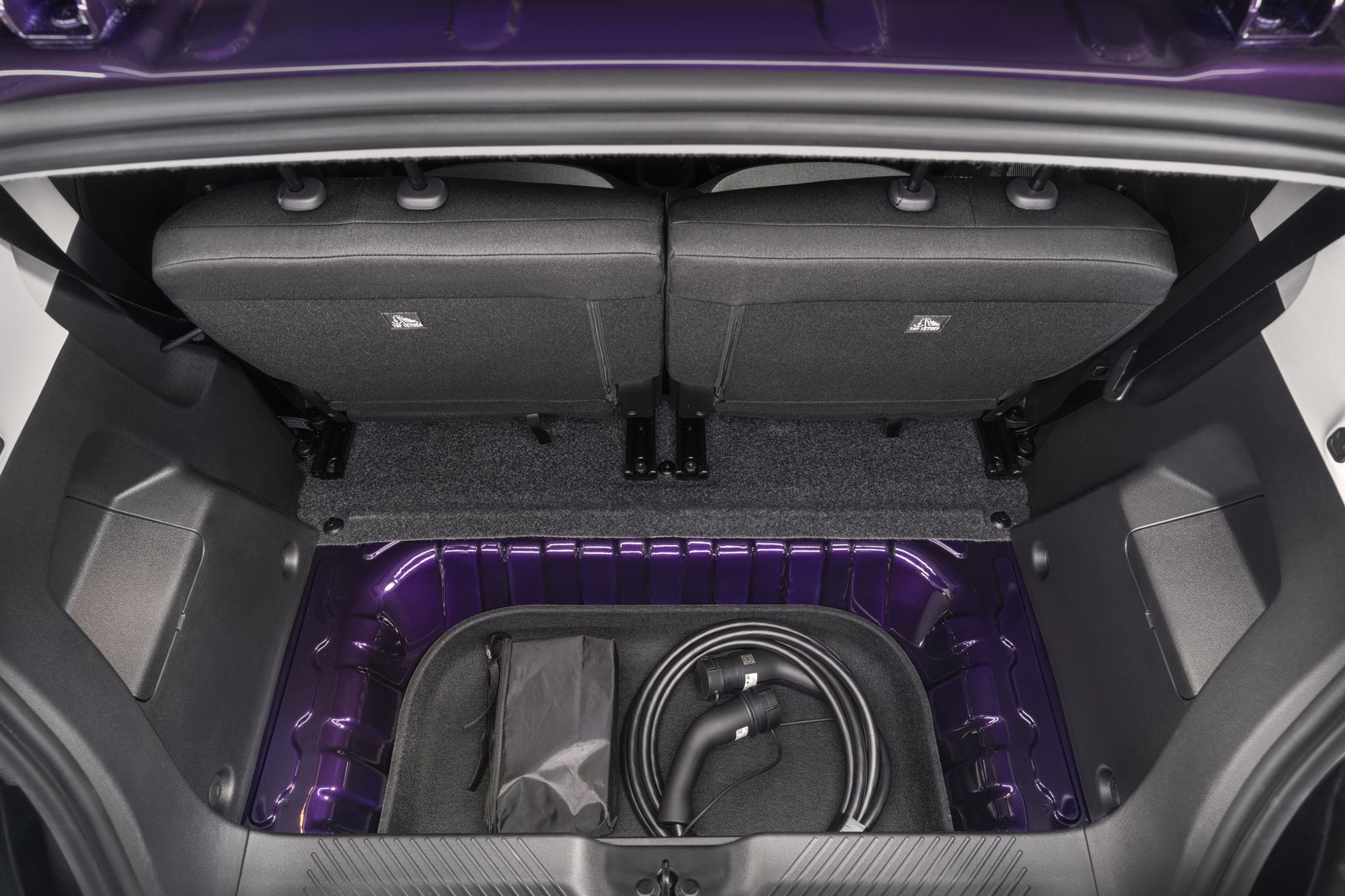 Photograph shows open rear cargo compartment of a purple vehicle, with split rear seatbacks raised. Lower storage well contains coiled charging cable and black pouch, surrounded by purple metal panels, molded side compartments, and textured trim.